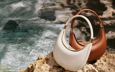 Two chic crescent-shaped handbags, one in classic white and the other in warm brown, rest on sun-kissed rocks by the ocean