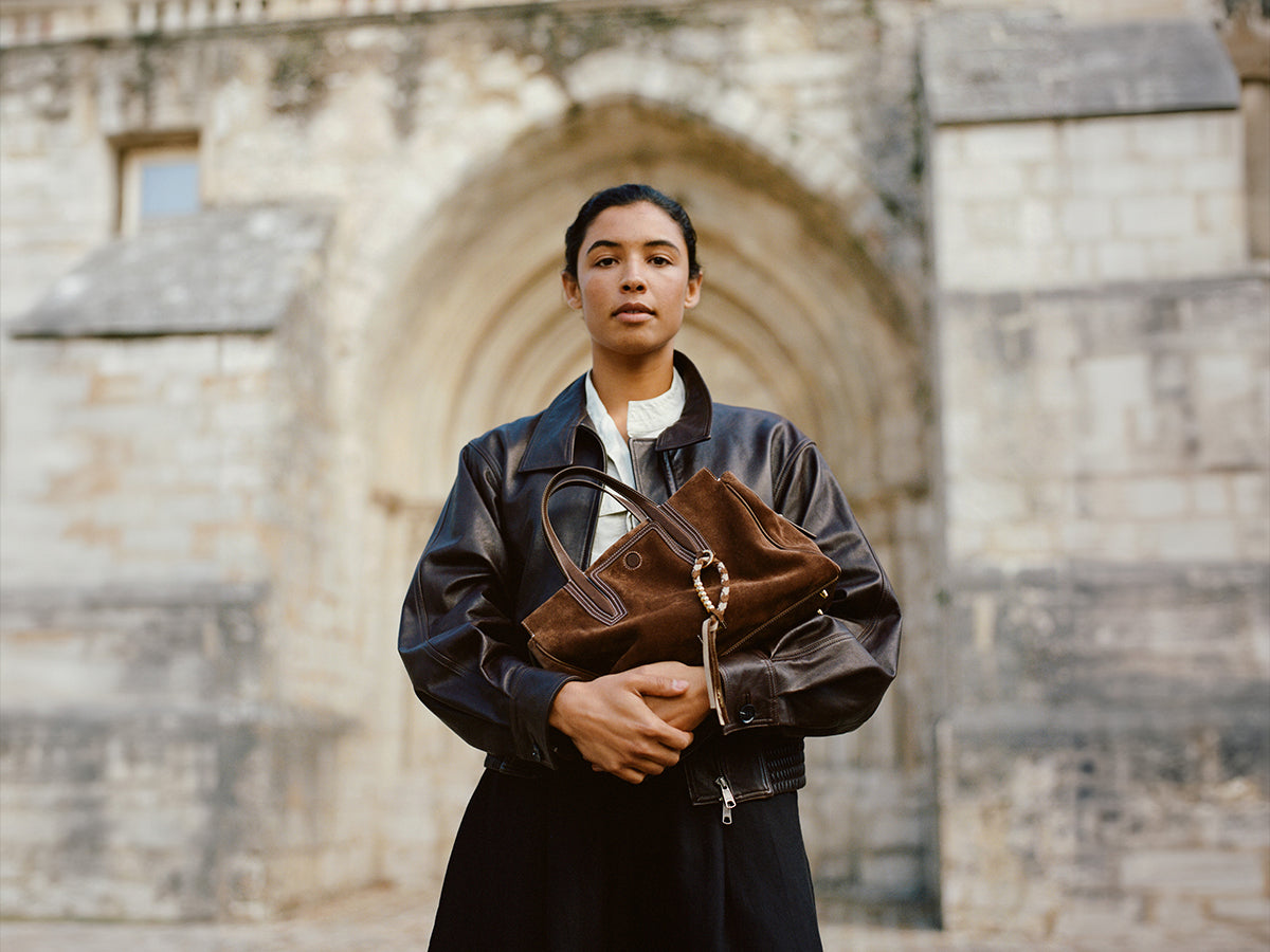 Person wearing a black jacket and skirt, holding a brown suede tote bag in front of a stone building