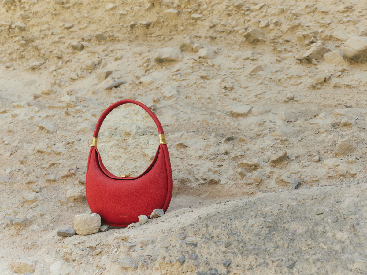 A stylish, crescent-shaped red handbag sits on a sandy