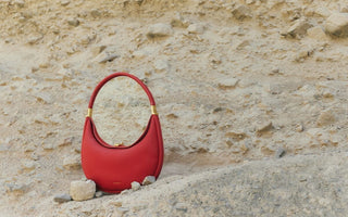 A stylish, crescent-shaped red handbag sits on a sandy