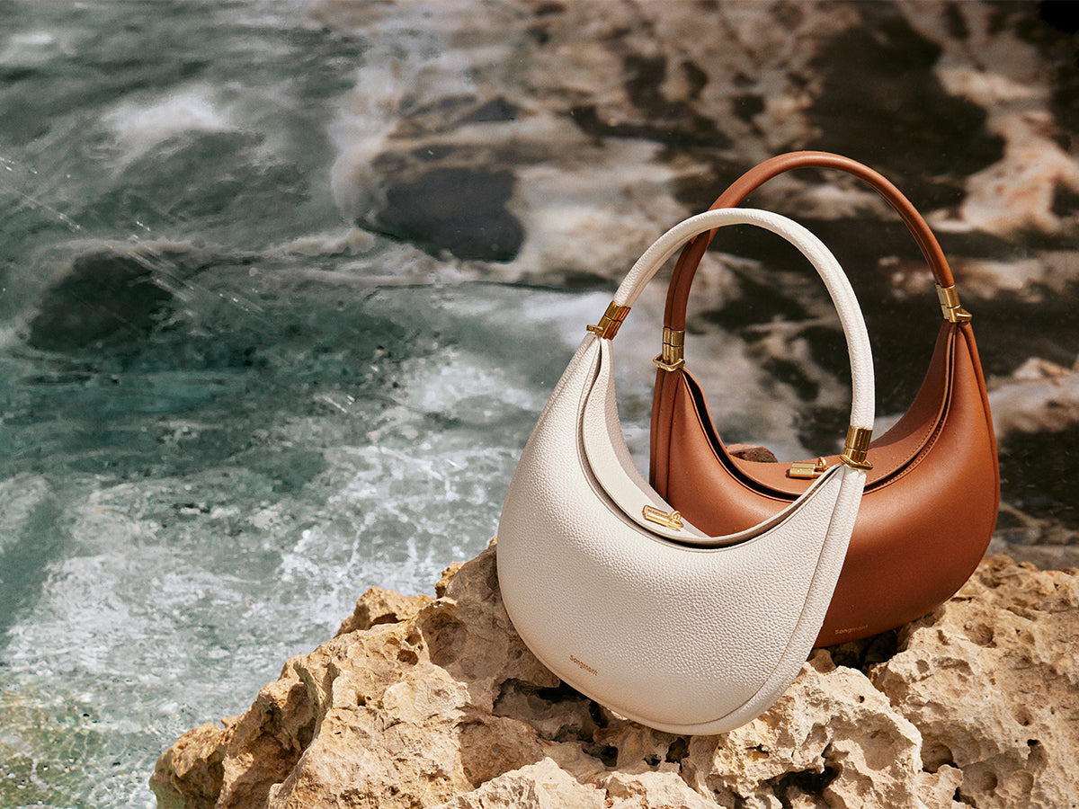 Two chic crescent-shaped handbags, one in classic white and the other in warm brown, rest on sun-kissed rocks by the ocean