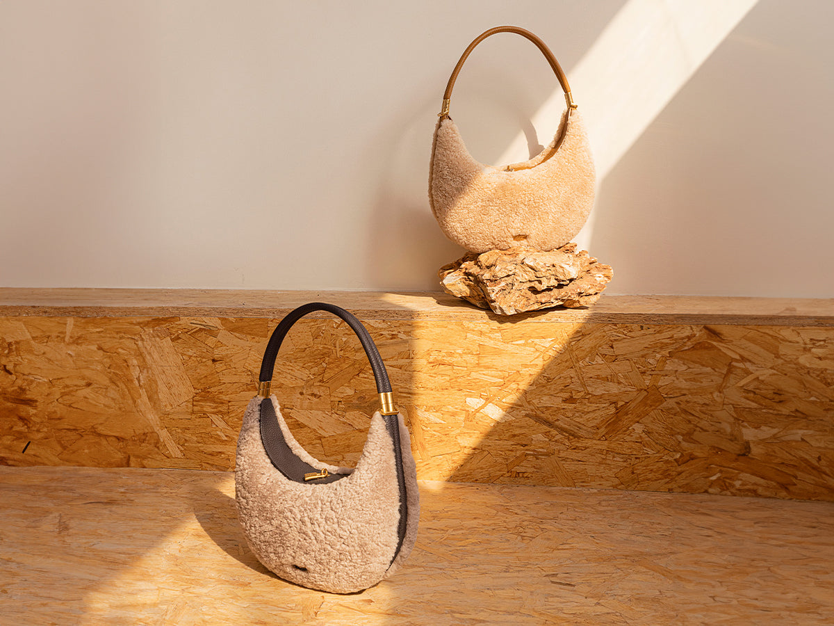 Beige shearling shoulder bag with a unique crescent moon shape and light brown leather handle, displayed on a textured wooden surface