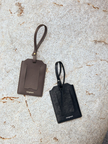 Songmont leather luggage tags in dark brown and black on a textured stone surface