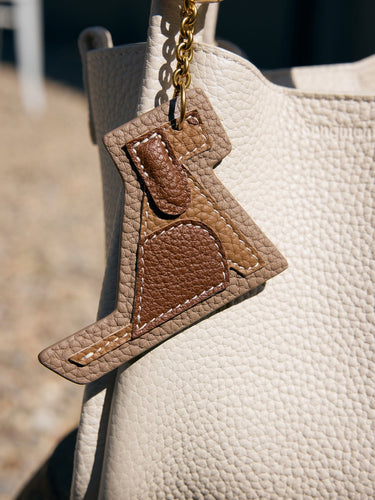 Songmont Puppy Charm accessory in tan and brown leather with white stitching on a beige handbag.