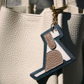 Songmont Puppy Charm accessory with navy blue trim and brown, white accents hanging from a cream leather bag.