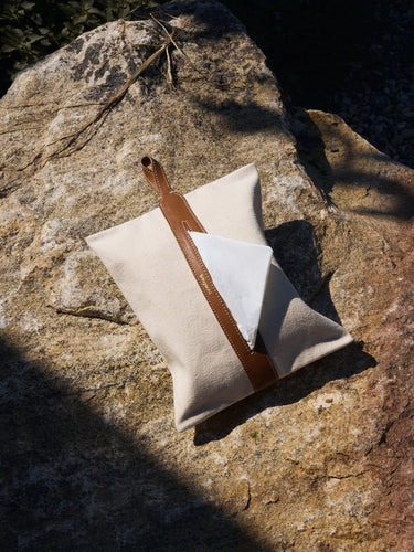 Songmont tissue box cover in natural canvas with a brown leather strap and gold logo, shown on a rock.