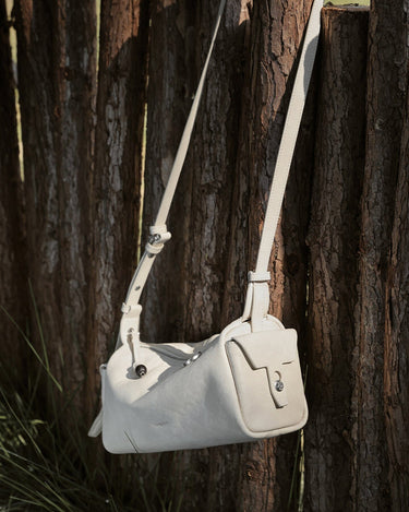 Mini Yore Camera Bag in Cloud color, made of vegetable-tanned leather, hanging on a wooden fence.