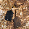 Two Songmont leather luggage tags, black and brown, with metal hardware on a textured rock.