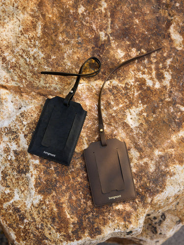 Two Songmont leather luggage tags, black and brown, with metal hardware on a textured rock.
