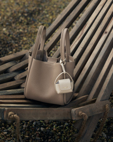 Songmont taupe full-grain leather tote bag with cream AirPods case attached, featuring magnetic closure and 'Songmont' text.
