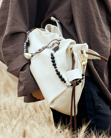 Cream leather Carefree Thread Charm bag with wood & grey beads, braided handles. Model in brown.
