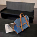 Faith Stone Charm handbag in denim blue with tan leather straps and gold buckle detail, placed next to a magazine.