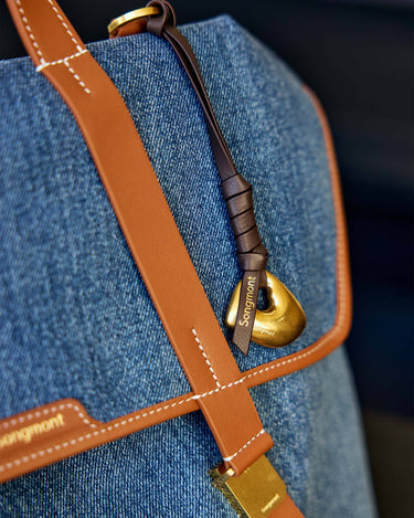 Faith Stone Charm bag in denim with tan leather trim, featuring a gold 'Songmont' logo charm and hardware.