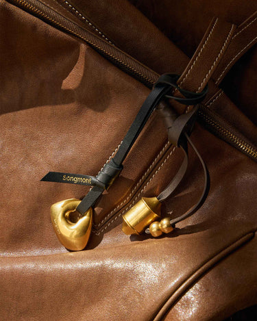 Faith Stone Charm accessory for a brown leather bag, featuring golden stone-like and cylindrical charms with beaded accents.