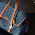 Denim Songmont bag with tan leather trim and straps, featuring a gold buckle.