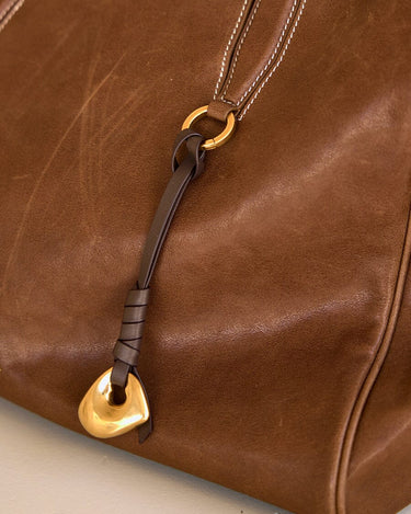 Faith Stone Charm detail on a brown leather bag, featuring a gold-tone charm and a dark brown leather strap.