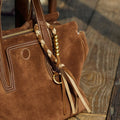 Brown suede Freedom Cord handbag with braided charm and tassels on a wooden surface