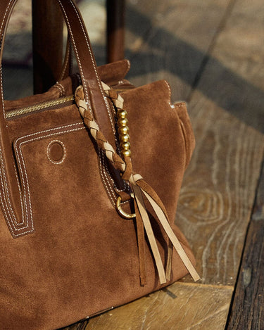 Brown suede Freedom Cord handbag with braided charm and tassels on a wooden surface