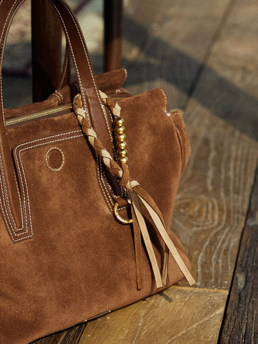 Brown suede Freedom Cord handbag with braided charm and tassels on a wooden surface