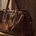 Brown suede Freedom Cord bag with leather trim and gold beaded tassel detail on a wooden surface.