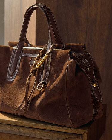 Brown suede Freedom Cord bag with leather trim and gold beaded tassel detail on a wooden surface.
