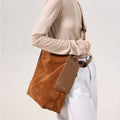 Large caramel suede Drippy tote bag with leather logo tag, worn on shoulder by a woman in beige top and light jeans.