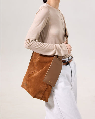 Large caramel suede Drippy tote bag with leather logo tag, worn on shoulder by a woman in beige top and light jeans.