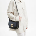Woman wearing a cream jacket and pants, carrying a black leather Large Reset Tofu Bag with a gold buckle.