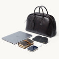 Dark brown full-grain nappa leather Yore bowling bag with white stitching, shown with a laptop, notebook, phone, and earbuds.