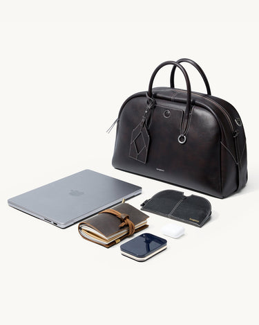 Dark brown full-grain nappa leather Yore bowling bag with white stitching, shown with a laptop, notebook, phone, and earbuds.