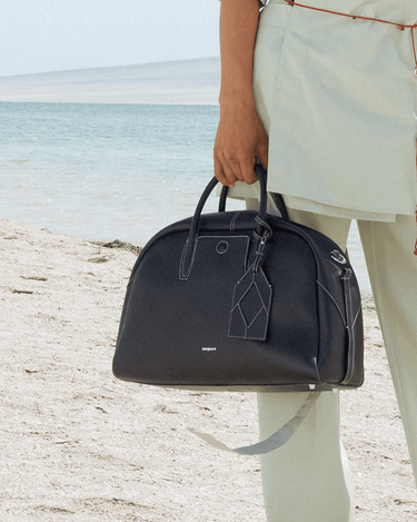Large navy full-grain leather Yore bowling bag with white stitching, held on a beach.