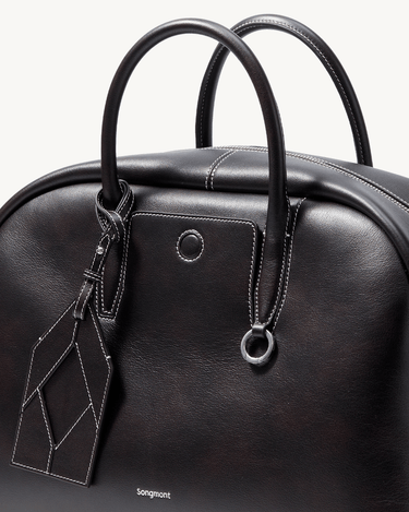 Large black Yore bowling bag made of full-grain Nappa leather with white stitching and a geometric luggage tag.