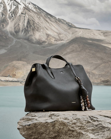 Large Yore Tote Bag in Onyx full-grain leather, with gold hardware and a beaded charm, set against a natural landscape.