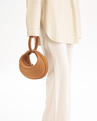 Tan pebbled leather Luna Bag with round handle and gold clasp, worn with a cream outfit.