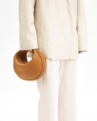 Pebbled tan leather Luna Bag with gold clasp, held by a person in cream outfit