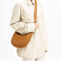 Luna Bag in tan pebble leather, worn by a woman in a cream oversized shirt and trousers.