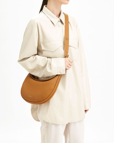 Luna Bag in tan pebble leather, worn by a woman in a cream oversized shirt and trousers.