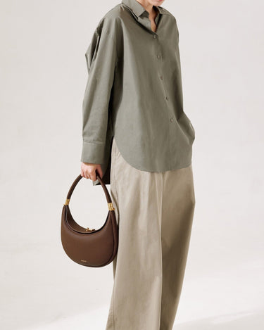 Chocolate pebbled leather Luna Bag with gold hardware, worn with a green shirt and tan pants.