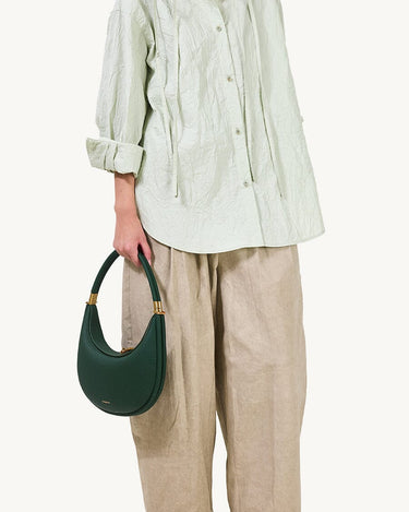 Emerald green pebbled leather Luna Bag with gold hardware, worn with light green blouse and beige pants.