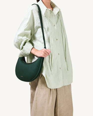 Luna Bag in Emerald green, textured leather with a crescent shape, worn by a model in an oversized light green shirt.