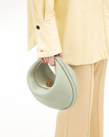 Light green pebbled leather Luna Bag with gold turn-lock closure, held with yellow shirt and beige pants.