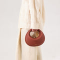Luna Bag in Oolong: A round, pebble-grain leather handbag with a gold clasp, held by a person in a beige linen outfit.