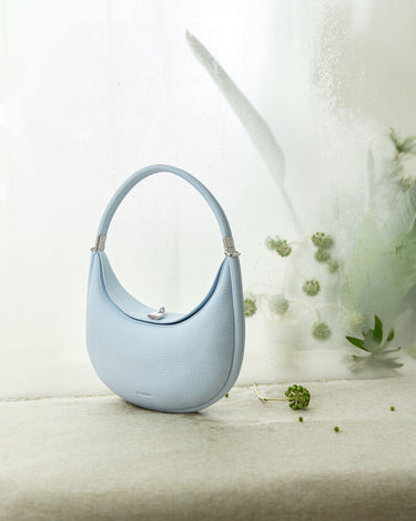 Light blue Luna Bag with pebble-grain leather texture, silver clasp, and curved handle on textured surface.