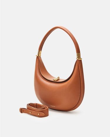 The Luna Bag in Eco Caramel vegan leather features a crescent shape, gold hardware clasp, and a detachable strap.