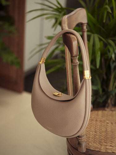 Taupe textured leather Luna Bag with gold hardware, hanging on a wooden chair.