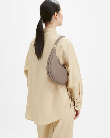 Taupe pebbled leather Luna Bag worn on the shoulder of a woman in a beige linen shirt.