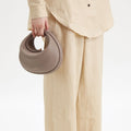 Taupe pebbled leather Luna Bag by Songmont with gold clasp, held by a person in beige linen clothing.