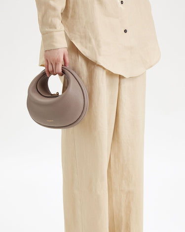 Taupe pebbled leather Luna Bag by Songmont with gold clasp, held by a person in beige linen clothing.