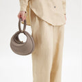 Small grey pebbled leather Luna Bag with curved handle and gold clasp, held by a person in beige linen outfit.