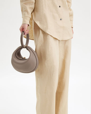 Small grey pebbled leather Luna Bag with curved handle and gold clasp, held by a person in beige linen outfit.